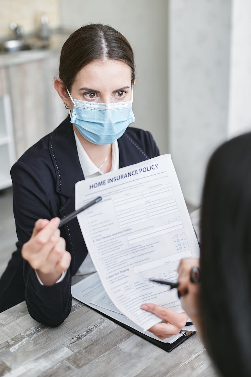 Professional woman wearing a face mask discussing a home insurance policy with a client at a desk.